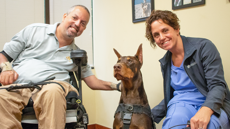 Owner and Service Dog Participating in the MedVet Service Dog Day Event Brown Doberman service dog sits between the veterinary ophthalmologist and his owner’s wheelchair after his free eye examination